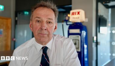 Duncan Lambert, wearing a white shirt and a blue tie sits in a room in front of an old Rix Petroleum petrol pump