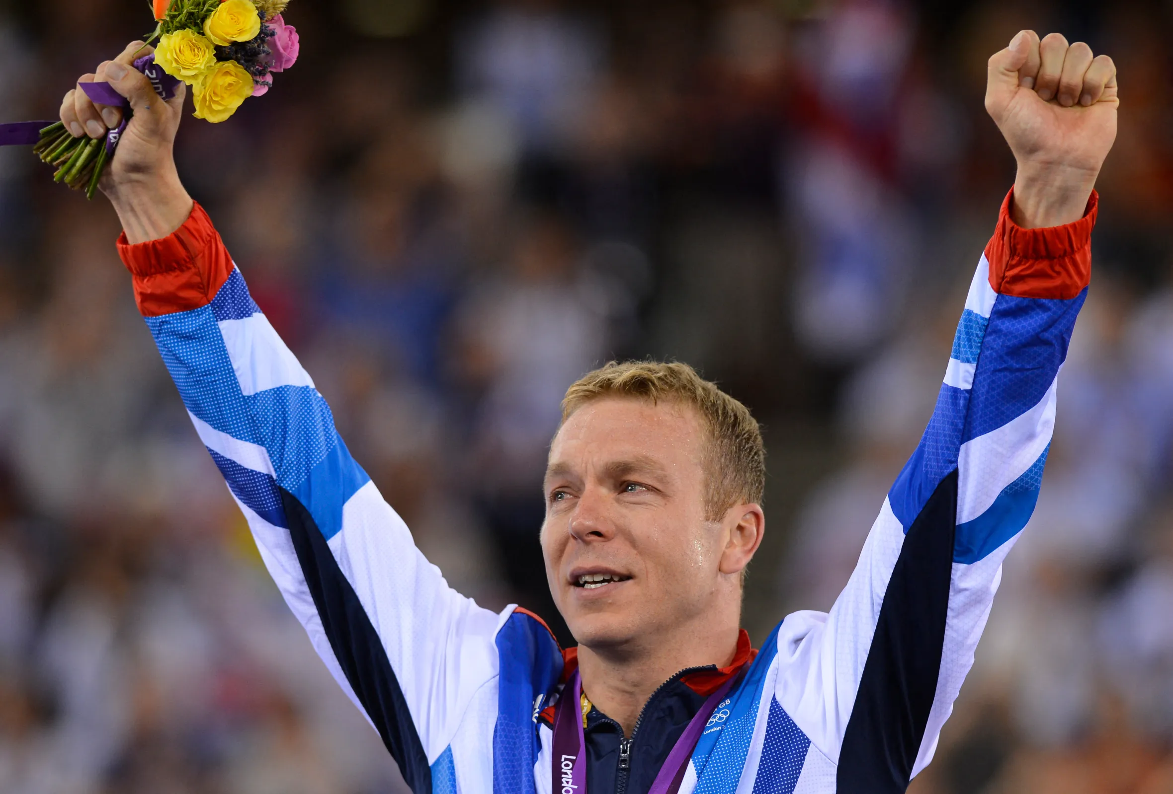 British cycling champion Chris Hoy on the podium with his arms raised, holding a bouquet of flowers in one hand.