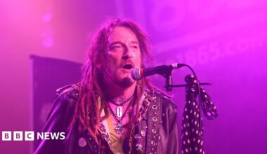 Ginger Wildheart performs with The Wildhearts at The 1865 in Southampton on 16 March 2025. He has long hair in dreadlocks, a short beard and moustache  and is wearing a leather jacket and an anarchy symbol and razor blade around his neck. He has musical notes tattooed along the base of his neck and there is a blue scarf with white polka dots tied to his microphone stand. He is singing into a microphone.