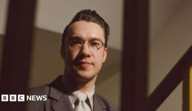 Mark Lamarr in glasses, a quiff, suit and tie in an official portrait photo taken in 1995