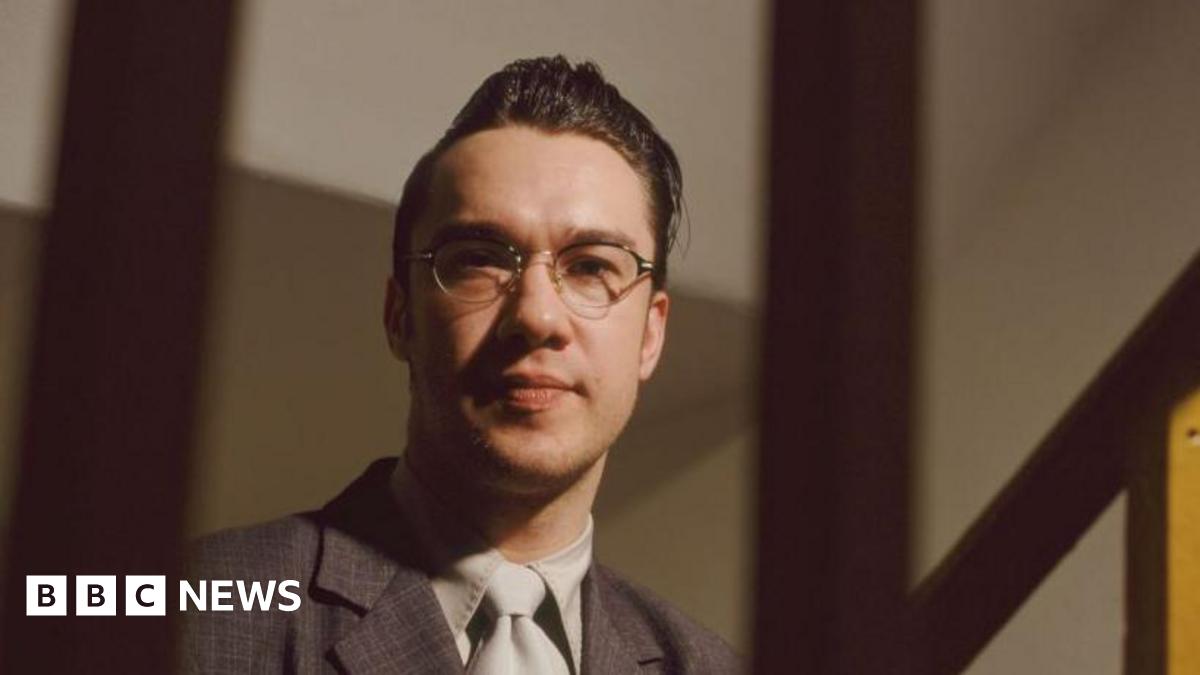 Mark Lamarr in glasses, a quiff, suit and tie in an official portrait photo taken in 1995