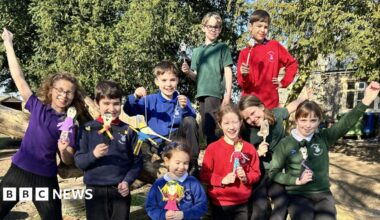 A group of children wearing different coloured school jumpers smile at the camera, all holding wooden spoons that have been decorated to look like their favourite book characters.