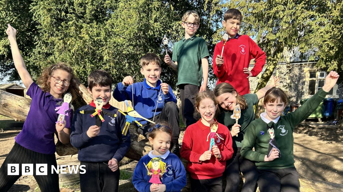 A group of children wearing different coloured school jumpers smile at the camera, all holding wooden spoons that have been decorated to look like their favourite book characters.
