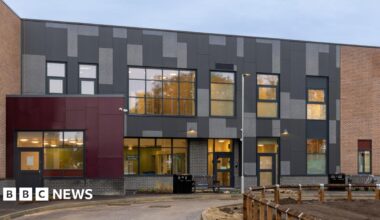 The exterior of the new mental health ward at Pilgrim Hospital in Boston. It is a multi-coloured building with large windows. It has a road in front of it and a muddy area surrounded by short wooden fencing.