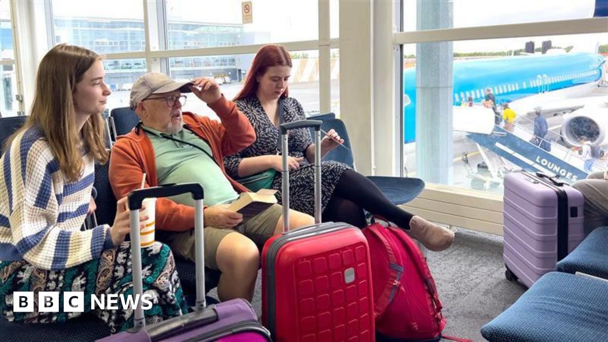 An older man in a baseball cap and an orange unzipped sweater sits between two younger women in an airport waiting area with small luggage trolleys. They're near a window through which a blue plane can be seen.