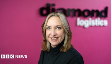 A woman with blonde hair. She is wearing a black jumper. The background behind her is pink.