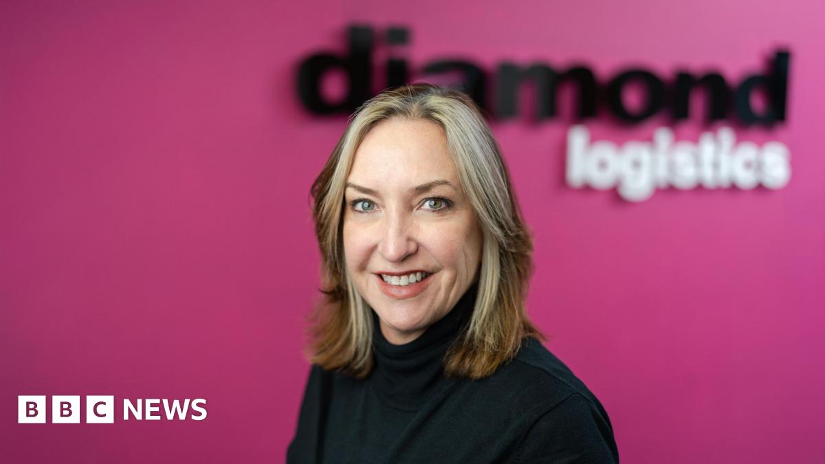 A woman with blonde hair. She is wearing a black jumper. The background behind her is pink.