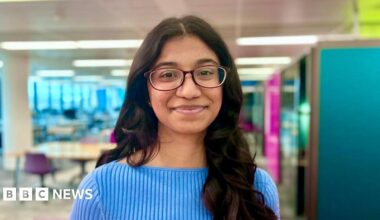 Bhuvana Chilukuri with long dark brown wavy hair and glasses wearing a pale blue ribbed top and standing with a university library environment behind her