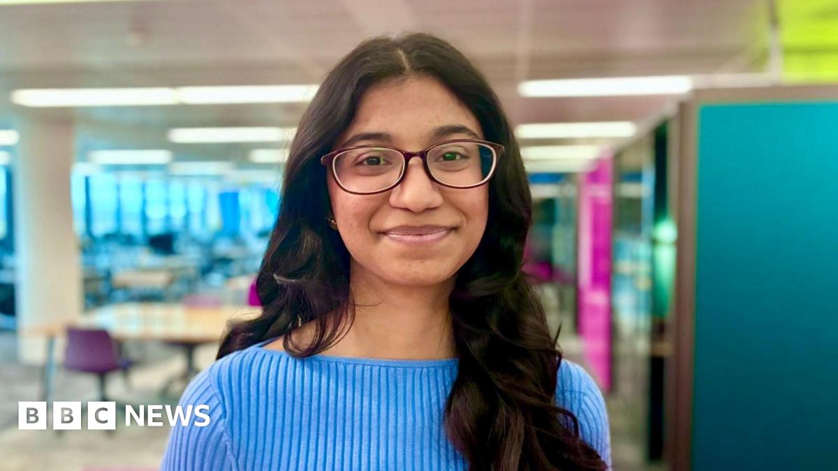 Bhuvana Chilukuri with long dark brown wavy hair and glasses wearing a pale blue ribbed top and standing with a university library environment behind her
