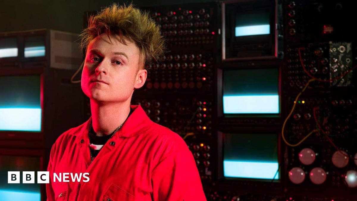 Look Mum No Computer, aka musician Sam Battle, stands in front of a bank of vintage synthesisers and TV monitors.