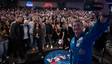 At right, a man in a blue flight suit with patches on it takes a selfie with his cell phone. He is in the photo, as well as panel members and the large audience. They are in an auditorium. In the far background, there is a NASA "meatball" insignia and the letters "JPL" on the wall.