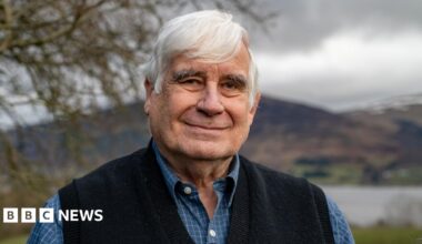 Mick North, a man with grey hair in a light blue shirt and dark blue tank top, stands in a garden. A loch, hills and tree can be seen behind him.