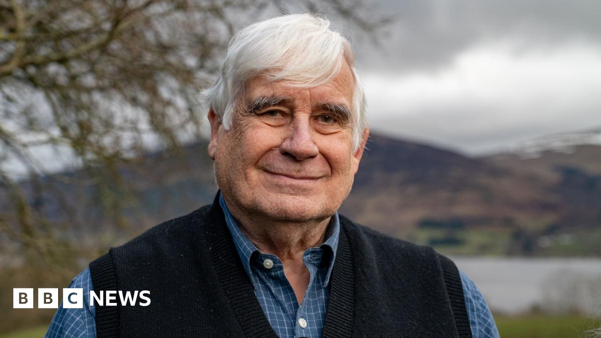 Mick North, a man with grey hair in a light blue shirt and dark blue tank top, stands in a garden. A loch, hills and tree can be seen behind him.