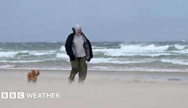 Man and his dog walking along a beach where sand is visibily lifted up with large waves in the background