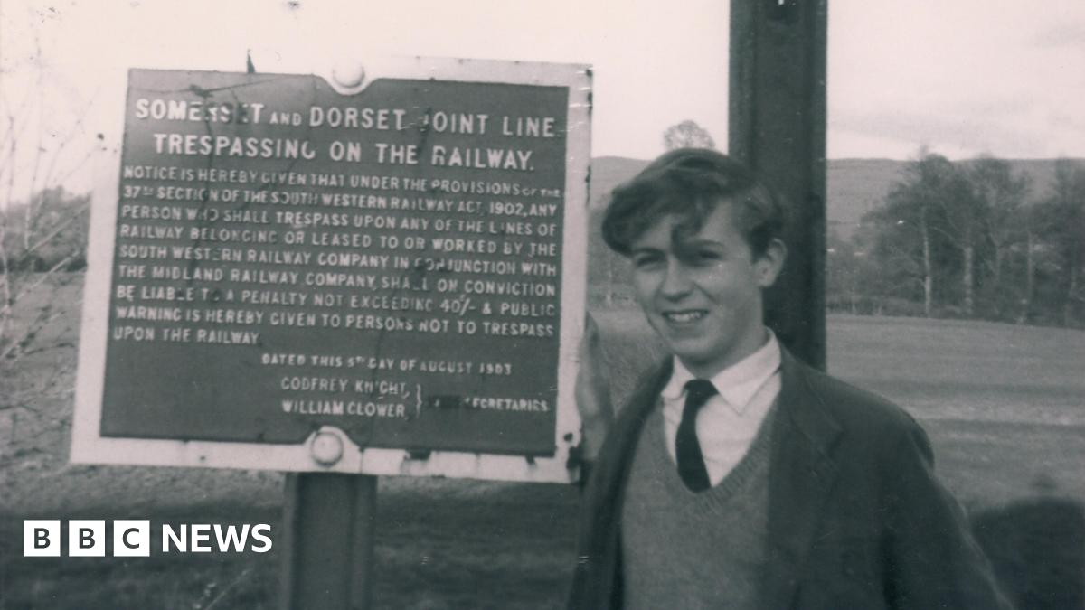 Black and white photo of Paul Antell as a teenager standing next to a sign which explains it is forbidden to trespass on the Somerset and Dorset line.