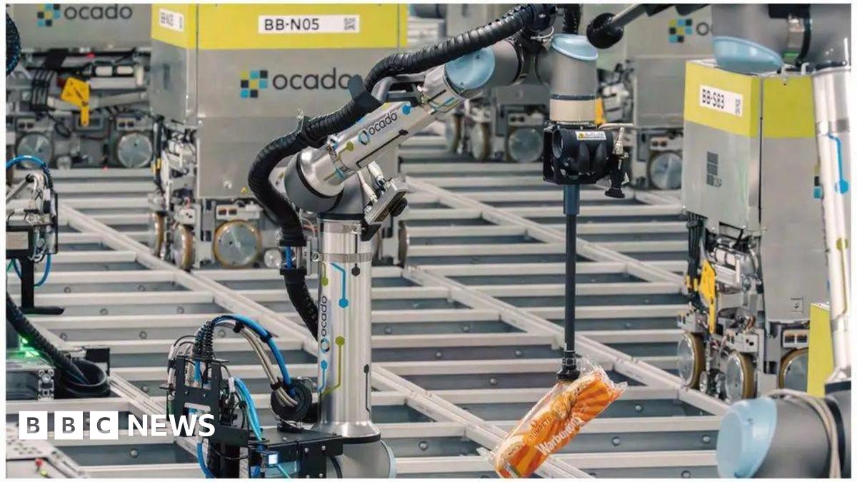 A series of silver robots move around on rails. In the foreground, one picks up Warburton crumpets with an arm