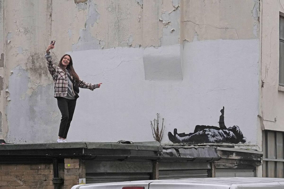 An individual in casual attire is positioned atop a building, their arms extended upward, with a large billboard behind them featuring a striking black-and-white image.