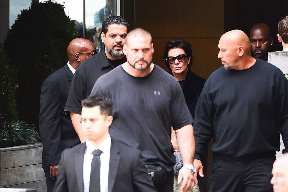 Several individuals dressed in formal attire are walking together in a corridor. Among them, two men are wearing black t-shirts, while another individual is dressed in a suit and tie. They appear to be moving towards an exit.