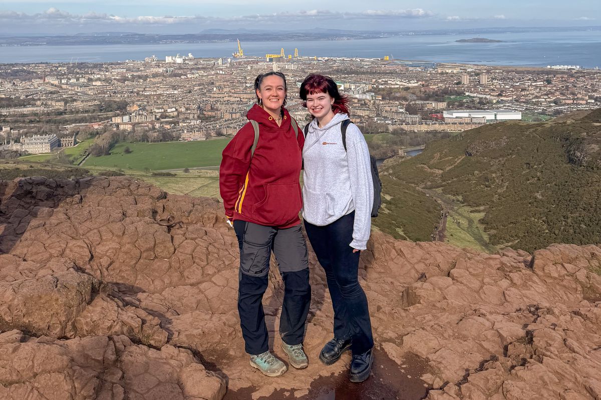 An image of two women standing on top of a hill with a city behind.