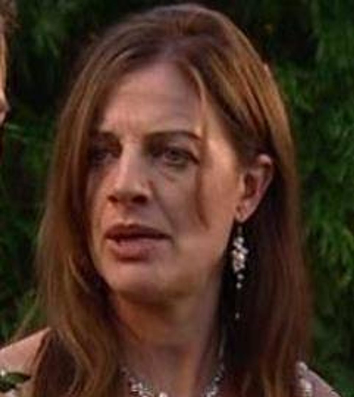 A close-up portrait of an individual with long, straight hair and a contemplative expression. The individual is adorned with elegant earrings and appears to be standing against a backdrop of foliage.
