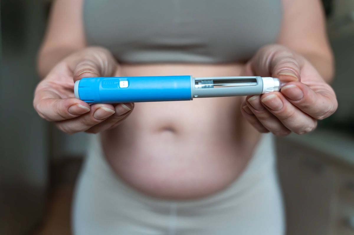 Close-up of a woman holding a GLP-1 injection pen in both hands with her bare abdomen visible in the background. The image illustrates weight management, hormone therapy, modern medical treatment, and body changes related to GLP-1 medications.