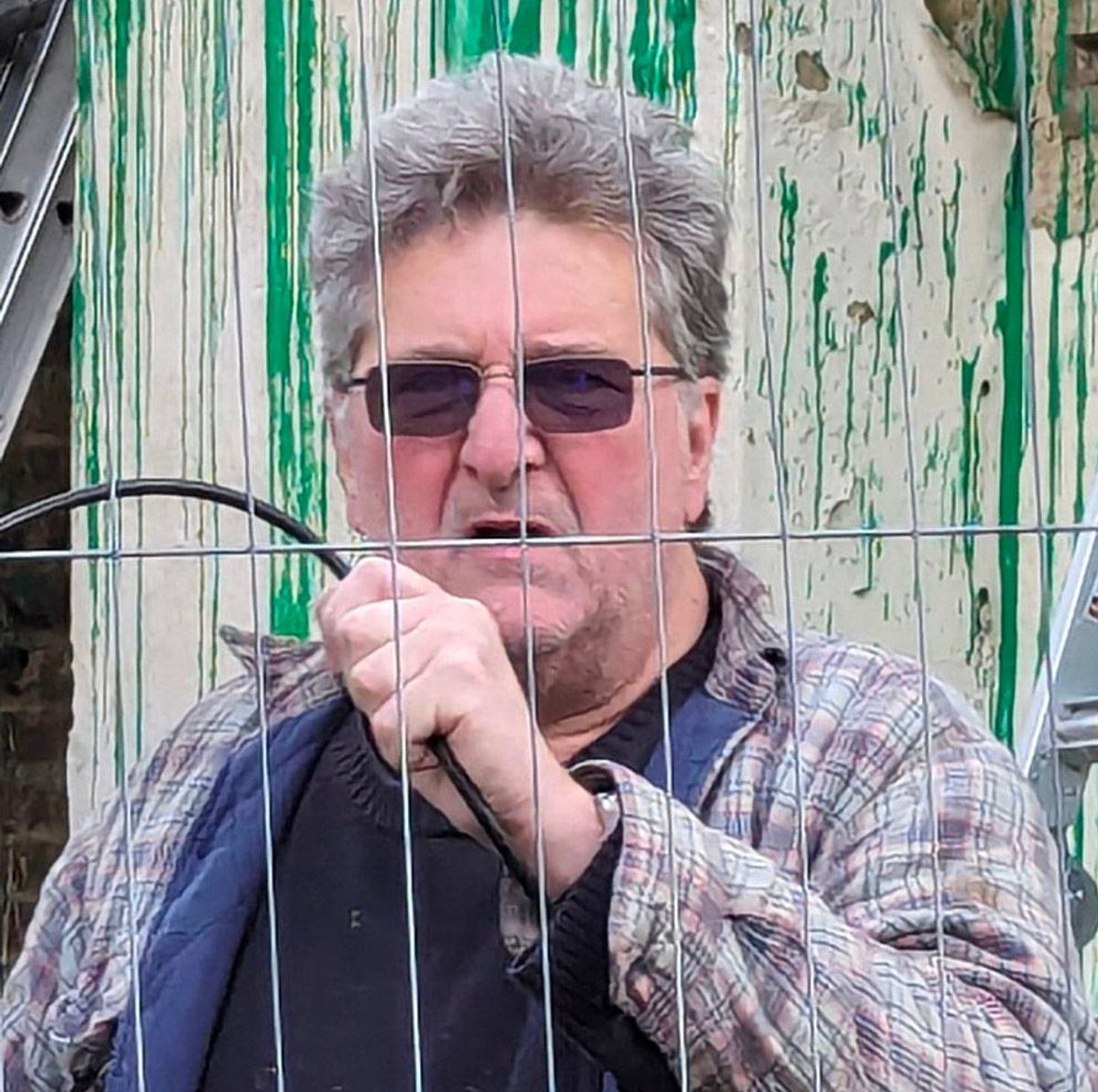 An older individual with gray hair, wearing sunglasses and a casual shirt, standing behind a wire mesh fence and holding a black object in their hand. The background features a green and white striped wall.