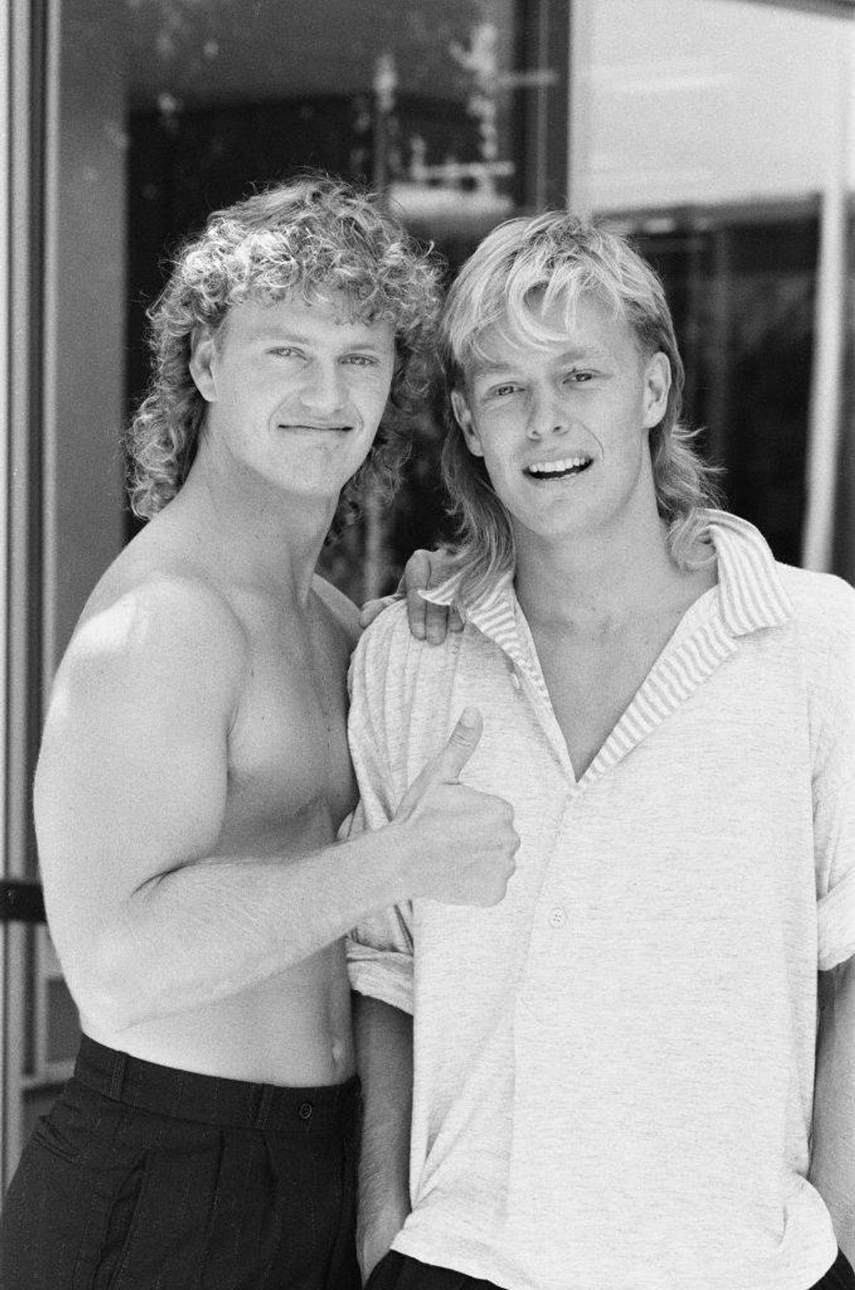 Two individuals, both with long, curly hair, are posing together for a photograph. The person on the left is shirtless, while the person on the right is wearing a white polo shirt. They both have cheerful expressions.