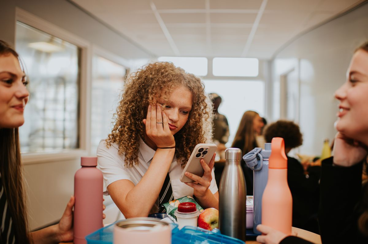 An antisocial school girl rudely excludes herself from a lively social conversation with peers in a school cafeteria. Her female friends talk animatedly as they eat lunch, whilst she stares at her phone, hand on chin, and looks bored and miserable.