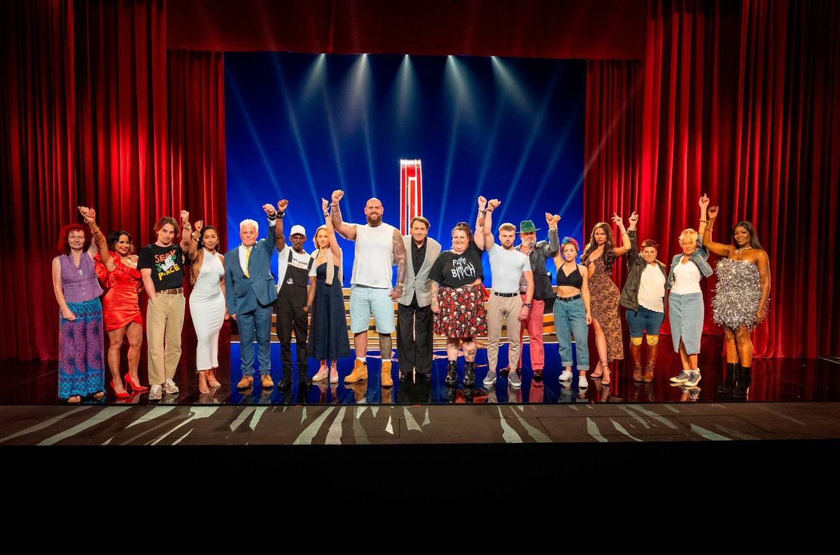 A group of individuals, dressed in a variety of attire, stand on a stage illuminated by spotlights, with a backdrop of red and blue curtains.