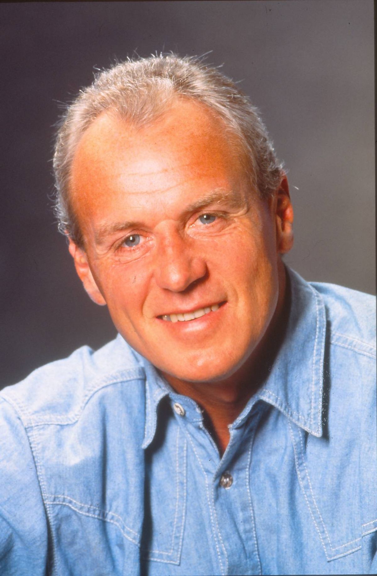 An individual with short gray hair and a blue shirt is smiling broadly, suggesting a state of contentment or happiness. The person’s facial expression and demeanor convey a sense of warmth and approachability.