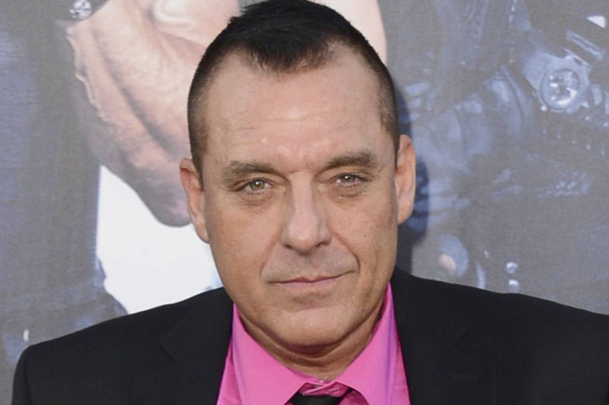 An individual dressed in a black suit with a pink shirt and a black tie is smiling and posing for a photograph against a blurred background featuring a hand.