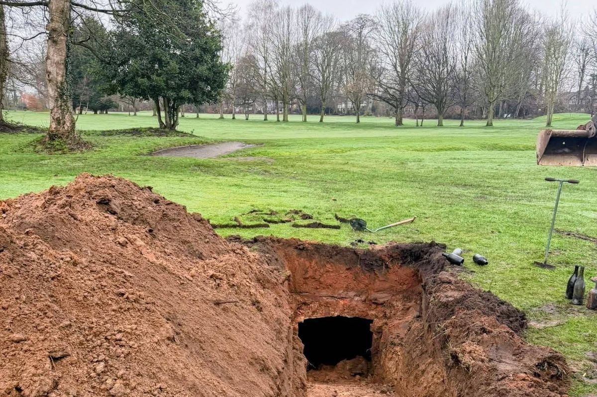 An aerial view of a construction site with a trench being dug into the ground, surrounded by a lush green field with trees and a clear blue sky in the background.