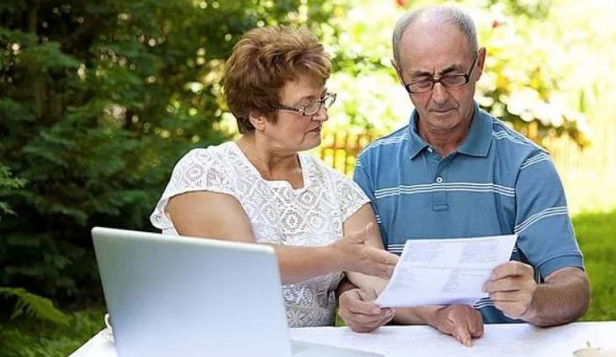 An older man and woman are sitting outside looking between a laptop screen and a piece of paper.