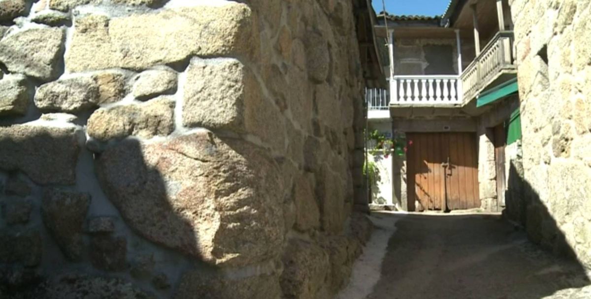 A narrow alleyway bordered by stone walls, leading to a building with a wooden entrance and a balcony adorned with white railings.