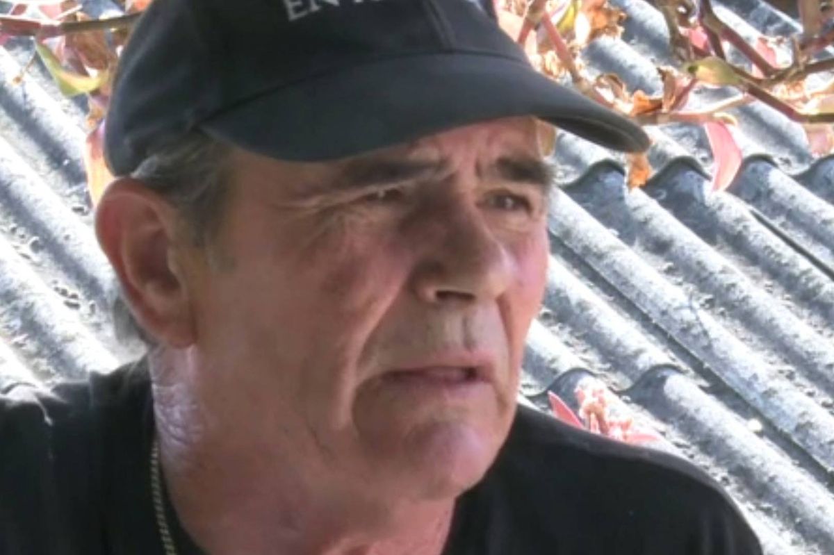 An individual wearing a black cap and a black shirt is seated and appears to be engaged in contemplation or focused attention. The background features a textured surface that resembles corrugated metal.