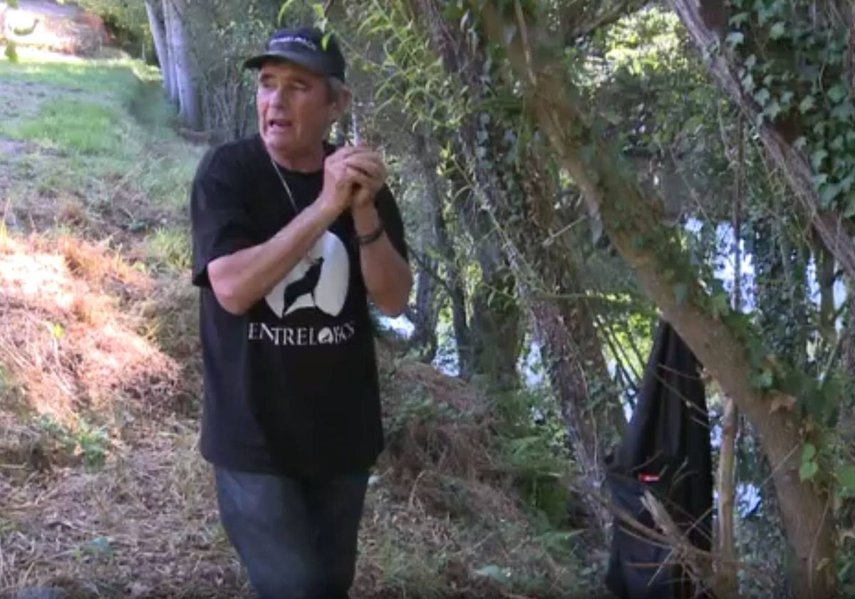 An individual dressed in a black t-shirt and jeans stands in a forested area, holding a small object, possibly a camera, and looking towards a body of water visible in the background.