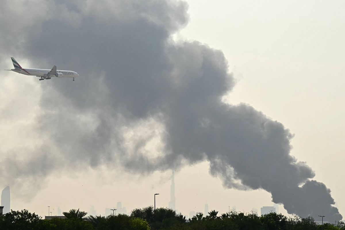 An Emirates aircraft flies past plumes of smoke from an ongoing fire near Dubai International Airport in Dubai on March 16, 2026. Missiles and drone attacks hit across the UAE, with a drone-related incident sparking a fuel tank fire near Dubai airport that disrupted travel, while a missile killed a civilian in Abu Dhabi. (Photo by AFP via Getty Images) /
