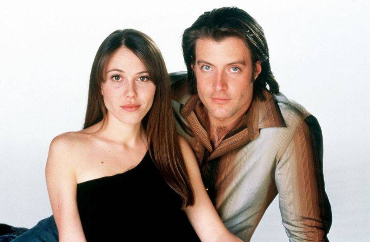 A man and a woman are sitting next to each other against a plain white background, posing for a photograph. The woman is wearing a black sleeveless dress, and the man is wearing a beige shirt with long hair. They both are looking directly at the camera.