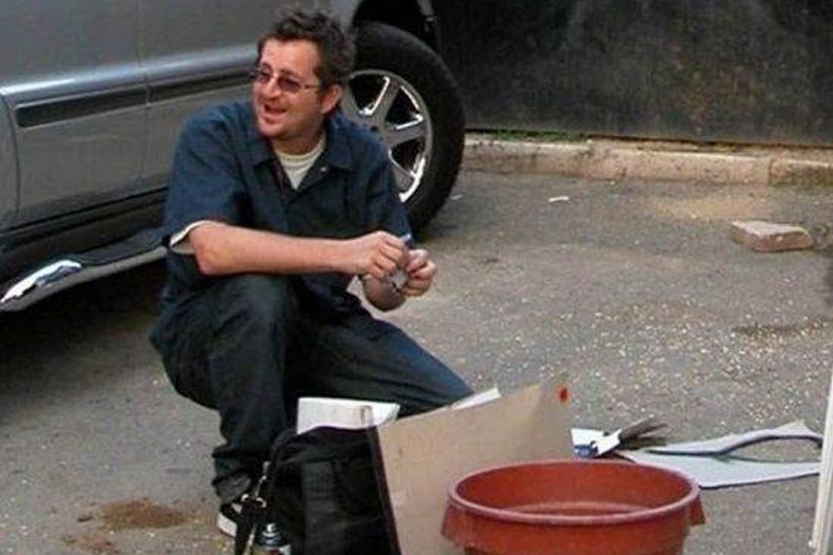 An individual is kneeling on a paved surface, engaged in an activity that involves a cardboard box and a red bucket. In the background, a vehicle is partially visible, with its front end positioned near a curb.