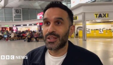 People with suitcases are hugged by loved ones in Stansted Airport's arrivals lounge, with the arrivals board visible behind them.