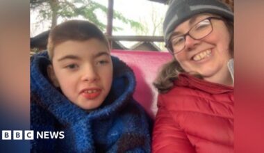 A selfie of a mum, who is wearing a red coat, grey beanie hat and black glasses, and her son, who is wearing a blue fleecy hoodie. They are sat at the back of a land train in a woodland setting.