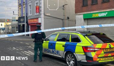A police officer stands with his back to the camera and is stood beside a police car. There is a police cordon closing off the street. In the distance there is the Lurgan police station.