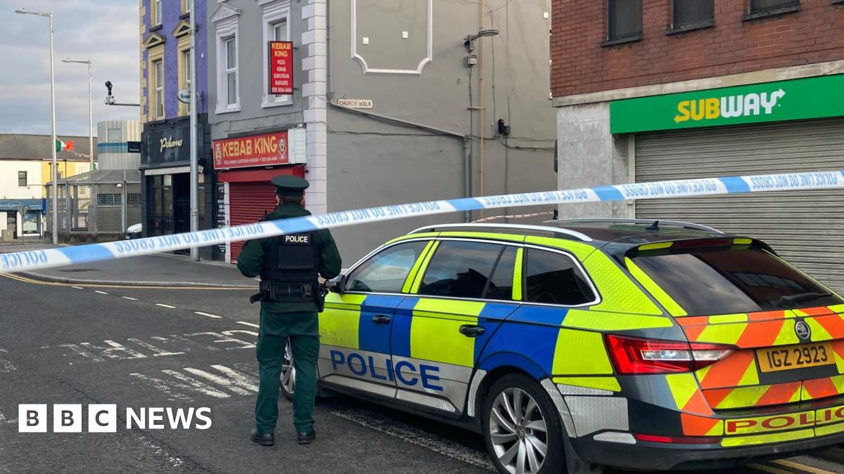 A police officer stands with his back to the camera and is stood beside a police car. There is a police cordon closing off the street. In the distance there is the Lurgan police station.