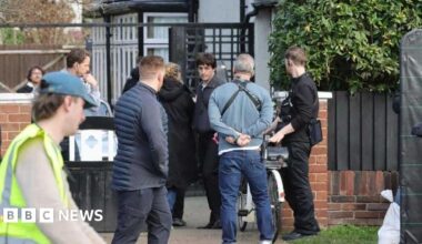 Paul Mescal is dressed in a blue jacket black trousers on a film set outside a beige coloured semi detached house.