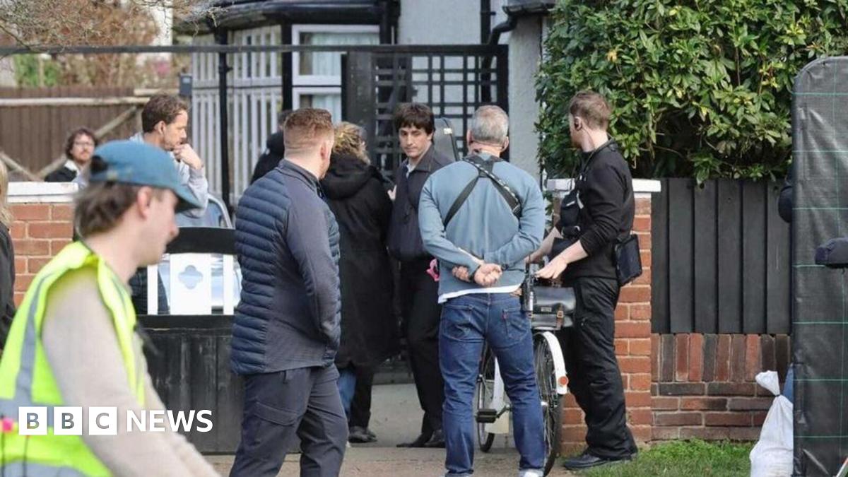 Paul Mescal is dressed in a blue jacket black trousers on a film set outside a beige coloured semi detached house.