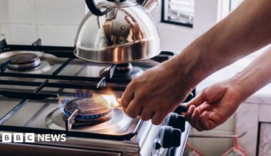 A person lights a gas stove using a match. There is a silver kettle sitting on one of the rings.