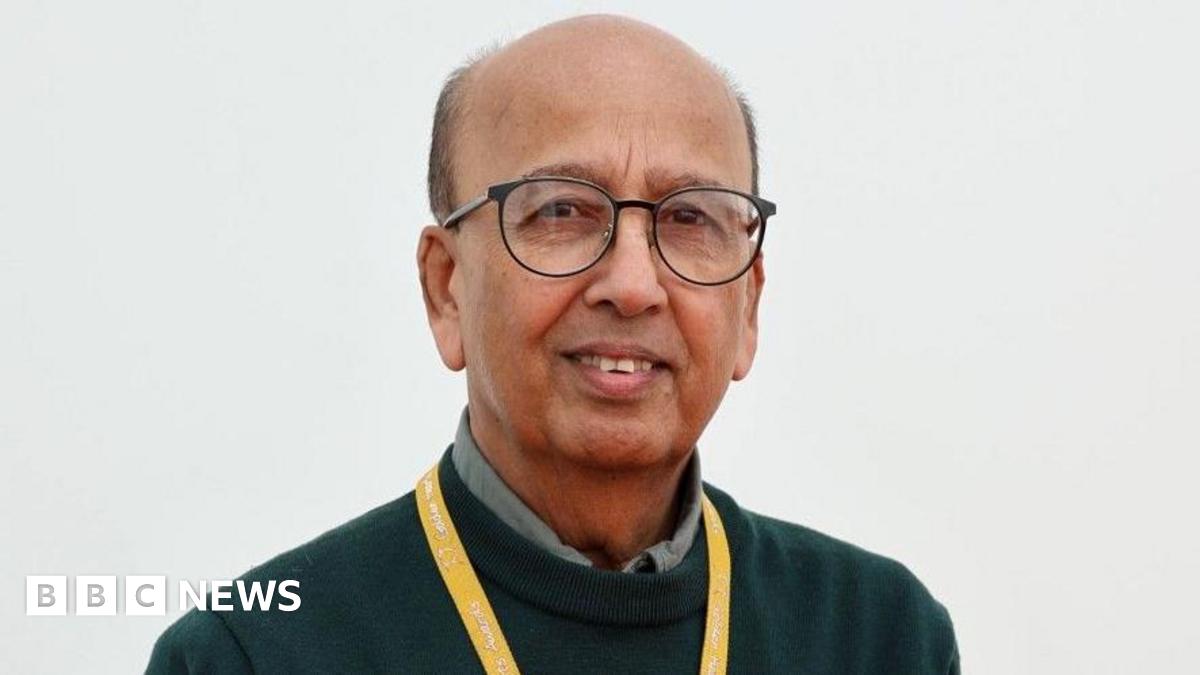 Prof Ahmed has heavily receding dark hair and is wearing black round spectacles on his face, a dark-green jumper over a sage-coloured shirt.