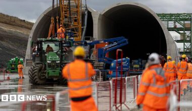 People in hard hats and high vis jackets in soft focus watch as a HS2 tunnel is built using a crane in front of them