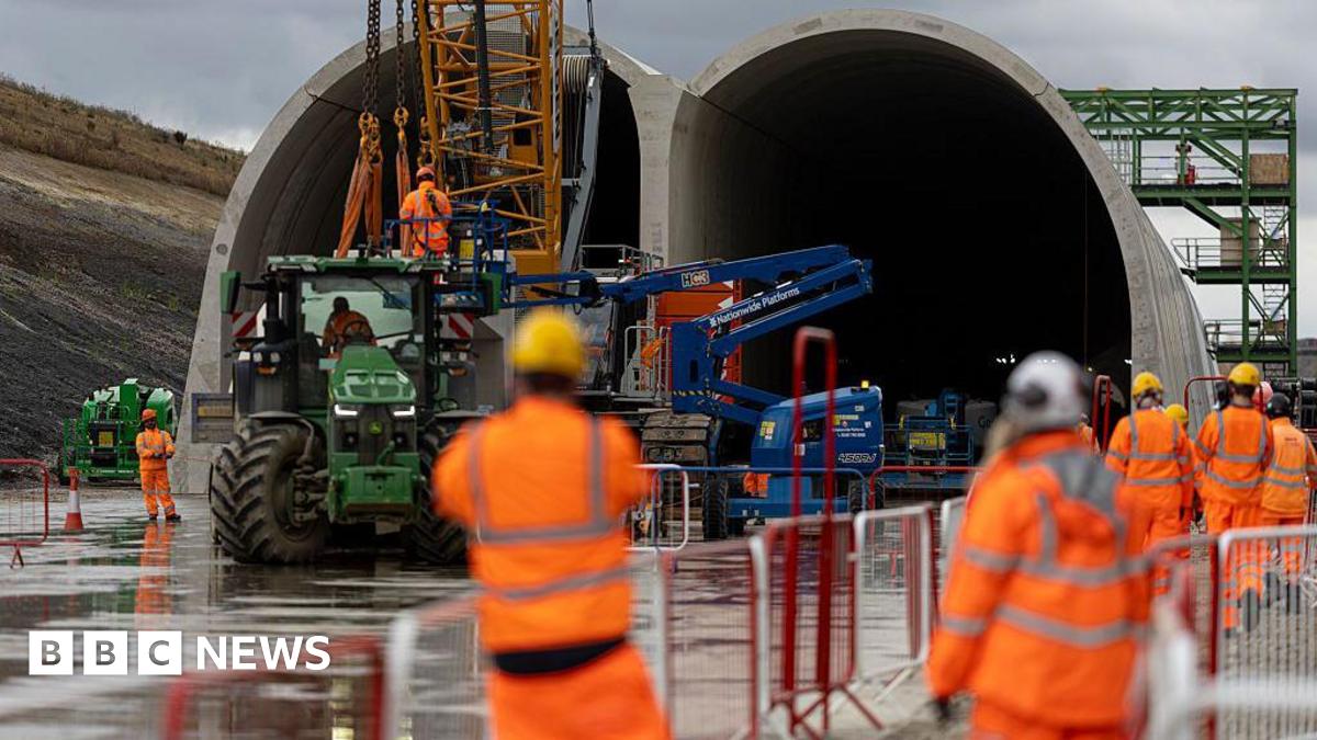 People in hard hats and high vis jackets in soft focus watch as a HS2 tunnel is built using a crane in front of them