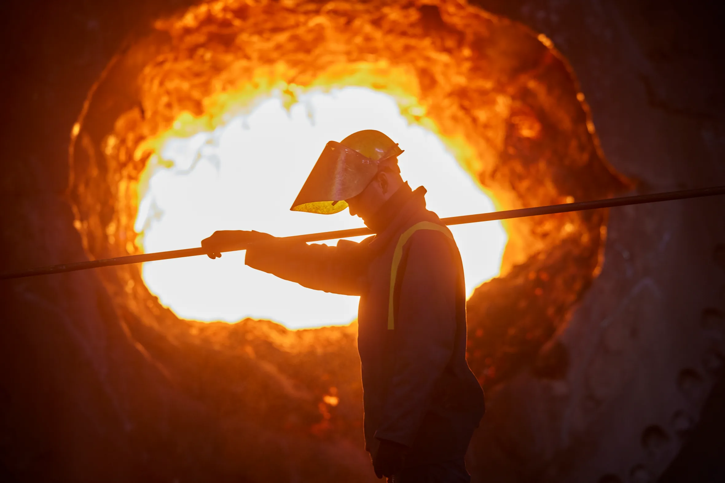 A worker wearing a hard hat and face shield works in front of a blast furnace.